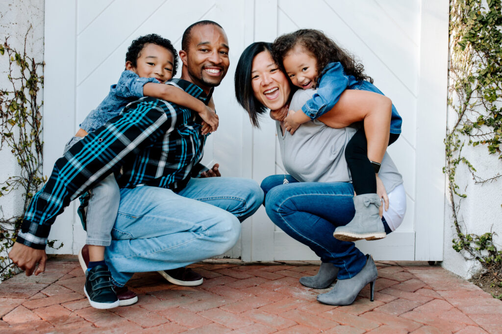 Asian mother and Black dad on brick porch with smiling mixed race children. Double white door is behind them.